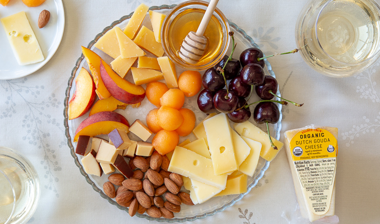 Cheese plate featuring three different Gouda cheeses, with cherries, sliced peaches, melon balls, almonds, and honey