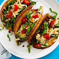 recipe: Soy Chorizo Tacos with shredded romaine, cheese, tomatoes, next to package of Trader Joe's Cauliflower Thins